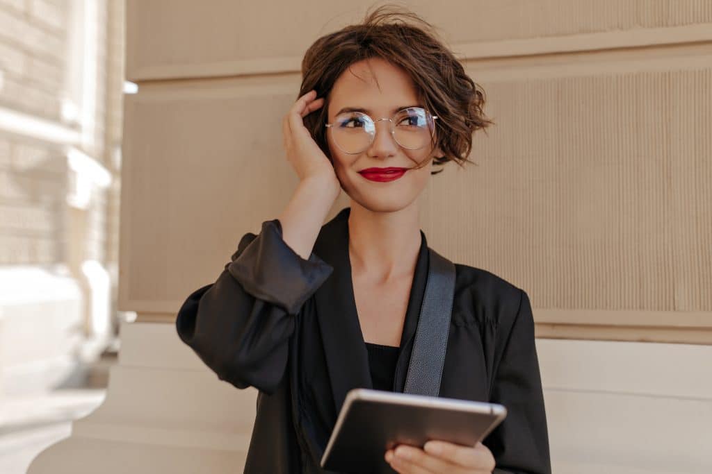 cute woman with short hair red lips smiling outside cheerful woman with red lipstick black clothes eyeglasses holds tablet outdoors